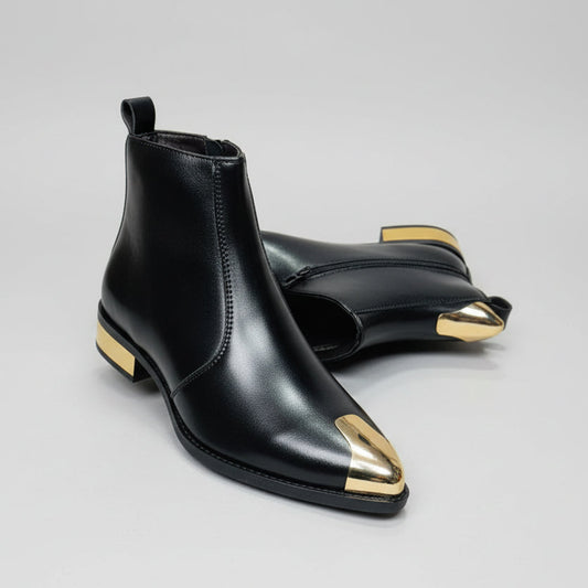 Black leather ankle boots with gold toes held by a hand, displayed on a wooden floor.